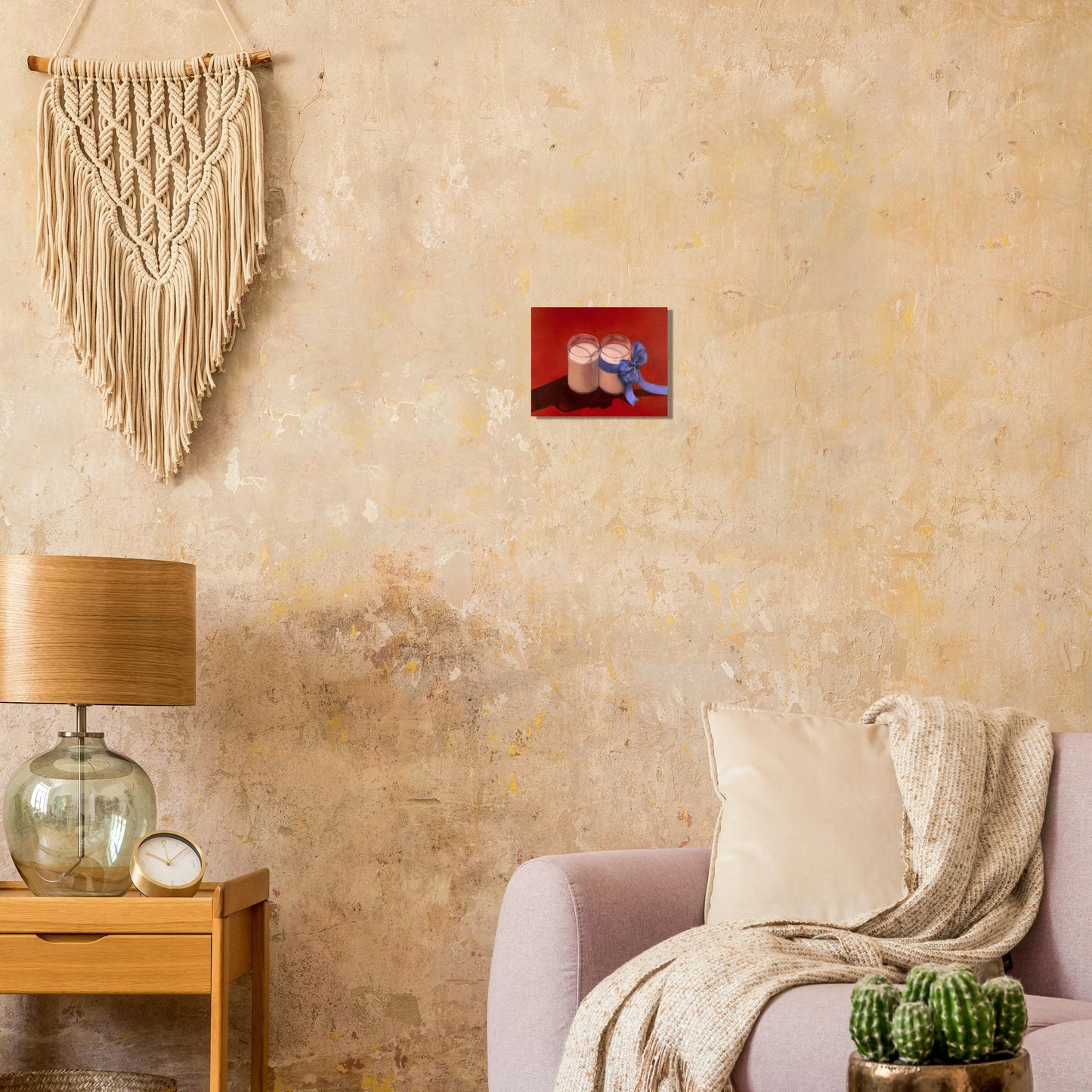 Jay Didyk's "Happy Holidays!" painting against a pale yellow stucco wall. A tan colored macrame hung on the wall off the to the left. Couch and oat-colored throw blanket, pot of cactus, table with lamp and clock on the bottom quadrant of the photo.